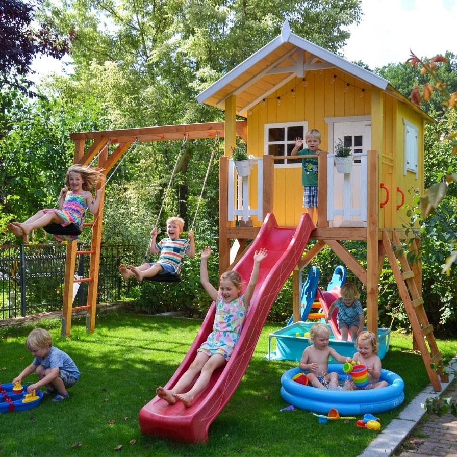Gelbes KASO Stelzenhaus Jonas L-A1 im Garten mit roter Rutsche, Doppelschaukel und spielenden Kindern auf der Wiese.