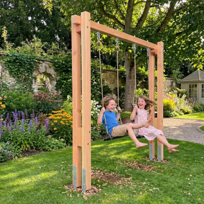 Kinder schaukeln glücklich auf einem massiven Douglasien-Holzschaukelgestell `Levi-1B-D` in einem blühenden Garten mit Steinmauer.