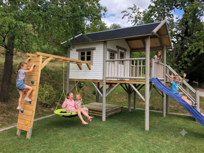 Seitenansicht des Stelzenhaus Hansson XXL-A6 mit Holz-Kletterwand inklusive bunten Klettersteinen, grüner Nestschaukel und zu öffnendem Seitenfenster.