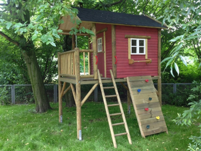Rotes Kinderspielhaus auf Stelzen im Garten mit Holzleiter und Kletterwand im Garten unter Bäumen.