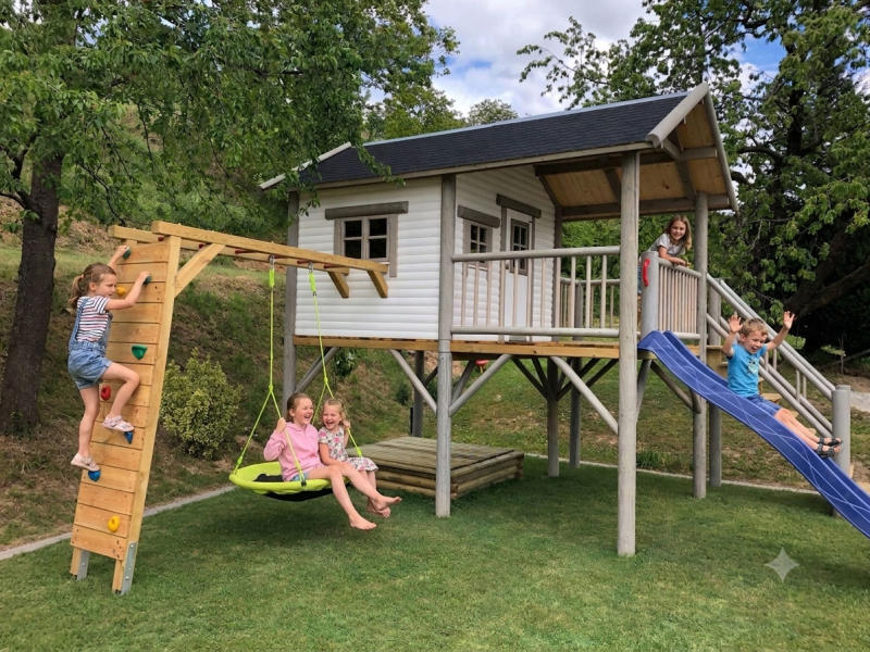 Seitenansicht des Stelzenhaus Hansson XXL-A6 mit Holz-Kletterwand inklusive bunten Klettersteinen, grüner Nestschaukel und zu öffnendem Seitenfenster.