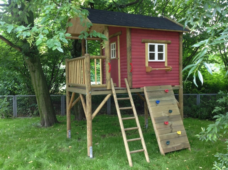 Rotes Kinderspielhaus auf Stelzen im Garten mit Holzleiter und Kletterwand im Garten unter Bäumen.