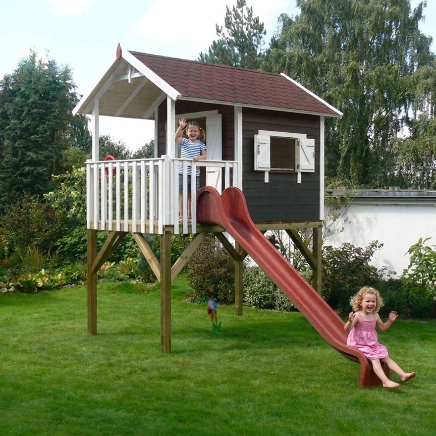 Stelzenhaus Janson XL von KASO aus dunkel gestrichenem Holz mit weißen Fensterläden, Veranda und roter Rutsche im Garten; ein Mädchen winkt von der Veranda, ein anderes sitzt am Ende der Rutsche.